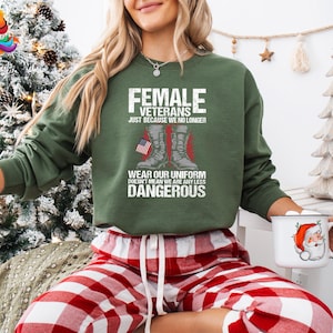 May include: Green sweatshirt with the text "Female Veterans Just Because We No Longer Wear Our Uniform Doesn't Mean We Are Any Less Dangerous". The text is in white and red. There are two boots with an American flag in the background.