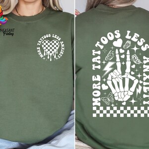Pu&ograve; includere: Felpa verde con una stampa grafica bianca che dice "Pi&ugrave; tatuaggi, meno ansia" con un cuore, un fulmine e un design a mano.