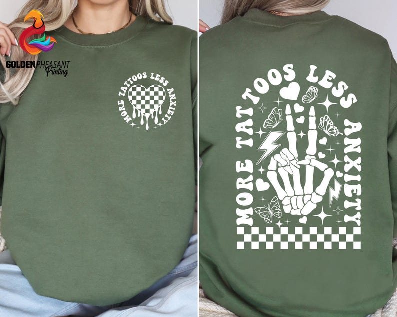Pu&ograve; includere: Felpa verde con una stampa grafica bianca che dice "Pi&ugrave; tatuaggi, meno ansia" con un cuore, un fulmine e un design a mano.