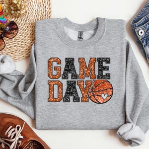 May include: A gray sweatshirt with the text "GAME DAY" in black and orange glitter. A basketball with a heart is in the middle of the text.