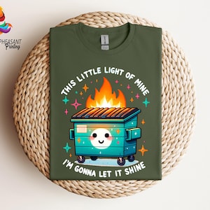 Pu&ograve; includere: Una maglietta verde con un'illustrazione di un cassonetto della spazzatura in fiamme in stile cartone animato. Il testo "This little light of mine, I'm gonna let it shine" &egrave; stampato sulla maglietta.