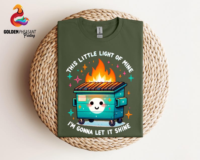 Pu&ograve; includere: Una maglietta verde con un'illustrazione di un cassonetto della spazzatura in fiamme in stile cartone animato. Il testo "This little light of mine, I'm gonna let it shine" &egrave; stampato sulla maglietta.