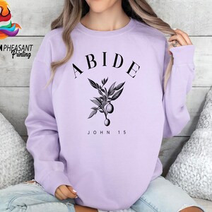 May include: A lavender sweatshirt with the word "ABIDE" in black text, a small black floral design, and the text "JOHN 15" in black text.