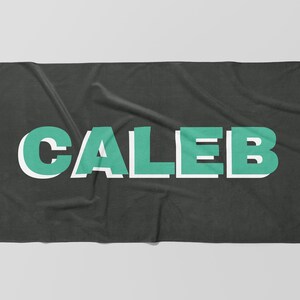May include: A dark gray blanket with the name "CALEB" in large, block letters. The letters are teal with a white outline, creating a 3D effect. The blanket is folded and has a soft, textured appearance.