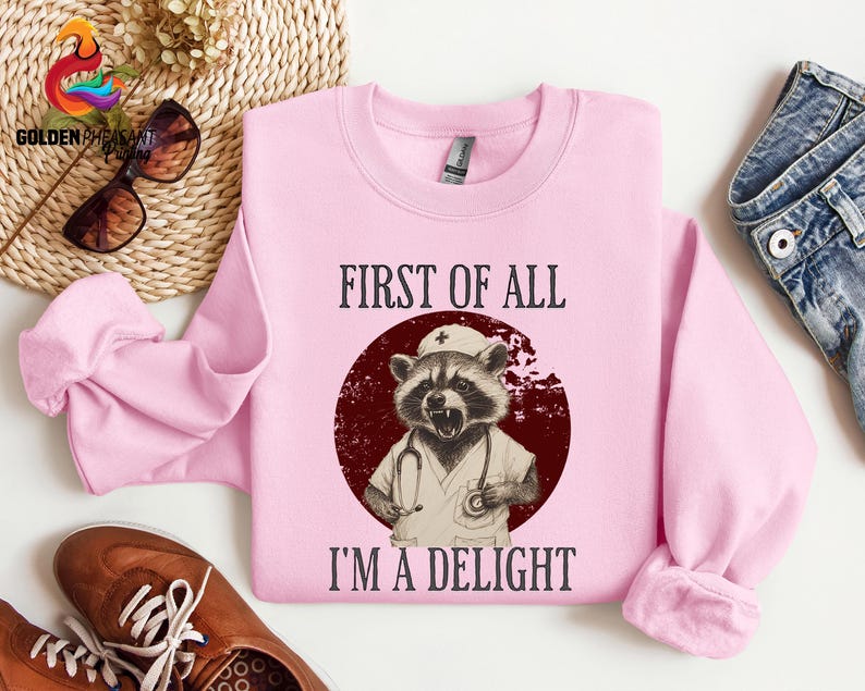 May include: Pink sweatshirt with a graphic of a raccoon wearing a nurse's uniform and a stethoscope. The text "First of all I'm a delight" is printed on the sweatshirt.