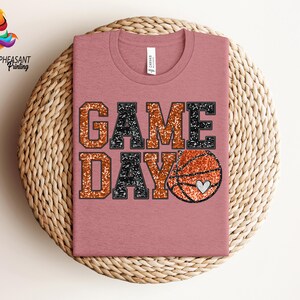 May include: A pink t-shirt with the words "GAME DAY" in black and orange glitter. A glittery orange basketball with a white heart is next to the words.