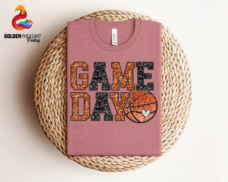 May include: A pink t-shirt with the words "GAME DAY" in black and orange glitter. A glittery orange basketball with a white heart is next to the words.