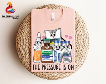 Pode incluir: Uma camiseta rosa com uma ilustração de desenho animado de suprimentos médicos, incluindo uma bolsa de fluido intravenoso, um frasco de epinefrina, um frasco de midazolam, um frasco de vasopressina e um frasco de fenilefrina. O texto "THE PRESSURE IS ON" é impresso abaixo da ilustração.