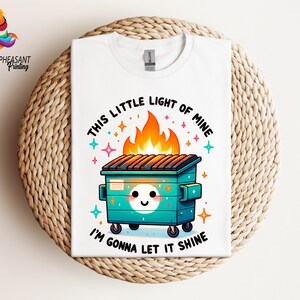 Pu&ograve; includere: Maglietta bianca con un cassonetto verde in fiamme con un viso sorridente e il testo "This little light of mine I'm gonna let it shine".