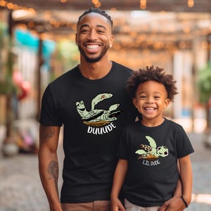 May include: Matching black t-shirts with a green sea turtle graphic. The larger shirt says "DUDE" and the smaller shirt says "LIL DUDE".