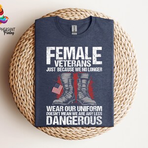May include: A navy blue t-shirt with a white graphic design featuring a pair of military boots and an American flag. The text on the shirt reads "Female Veterans Just Because We No Longer Wear Our Uniform Doesn't Mean We Are Any Less Dangerous".
