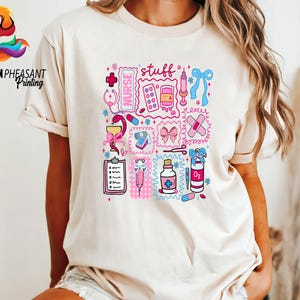 May include: Beige t-shirt featuring a colorful graphic of medical supplies and the word "stuff". The design includes a stethoscope, pills, a syringe, a bandage, and a bow. The shirt has short sleeves and a crew neck.