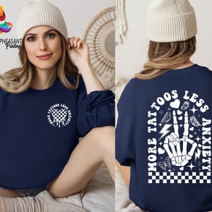 Pu&ograve; includere: Felpa blu navy con un design grafico bianco che presenta una mano scheletrica con un cuore, fulmini, farfalle e il testo "Pi&ugrave; tatuaggi, meno ansia".
