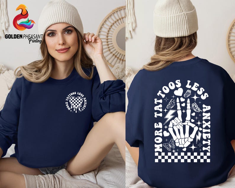 Pu&ograve; includere: Felpa blu navy con un design grafico bianco che presenta una mano scheletrica con un cuore, fulmini, farfalle e il testo "Pi&ugrave; tatuaggi, meno ansia".