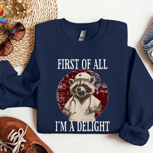 May include: Navy blue sweatshirt with a raccoon wearing a nurse's uniform and a stethoscope. The text "First of all I'm a delight" is printed on the sweatshirt.