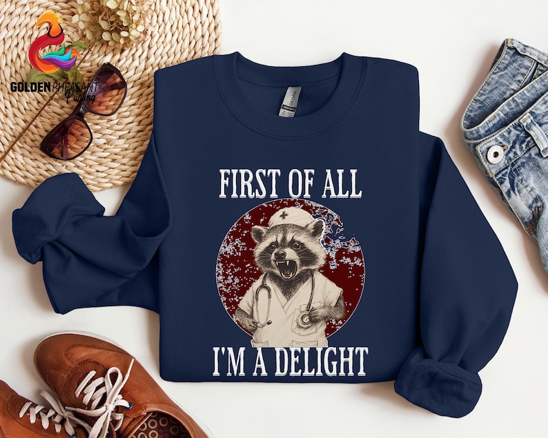 May include: Navy blue sweatshirt with a raccoon wearing a nurse's uniform and a stethoscope. The text "First of all I'm a delight" is printed on the sweatshirt.