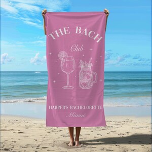 May include: A pink beach towel featuring white line art of a wine glass and a mason jar cocktail. The text reads THE BACH CLUB, HARPER'S BACHELORETTE, and Miami, accented with small star symbols across the center of the fabric.