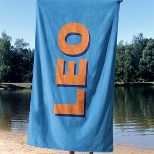 May include: A blue beach towel with the word "LEO" in large orange letters. The towel is held up against a sunny sky, with a lake and trees in the background. The towel is made of a soft material.