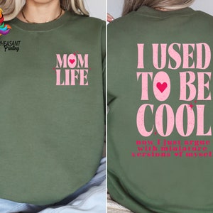 May include: A green sweatshirt with pink glitter text. The front of the sweatshirt says "MOM LIFE" with a pink heart. The back of the sweatshirt says "I USED TO BE COOL now I just argue with miniature versions of myself".
