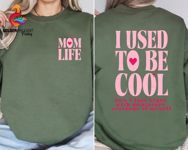 May include: A green sweatshirt with pink glitter text. The front of the sweatshirt says "MOM LIFE" with a pink heart. The back of the sweatshirt says "I USED TO BE COOL now I just argue with miniature versions of myself".