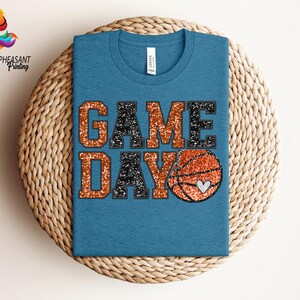 May include: A blue t-shirt with the words "GAME DAY" in black and orange glitter. A basketball with a heart is in the middle of the text.