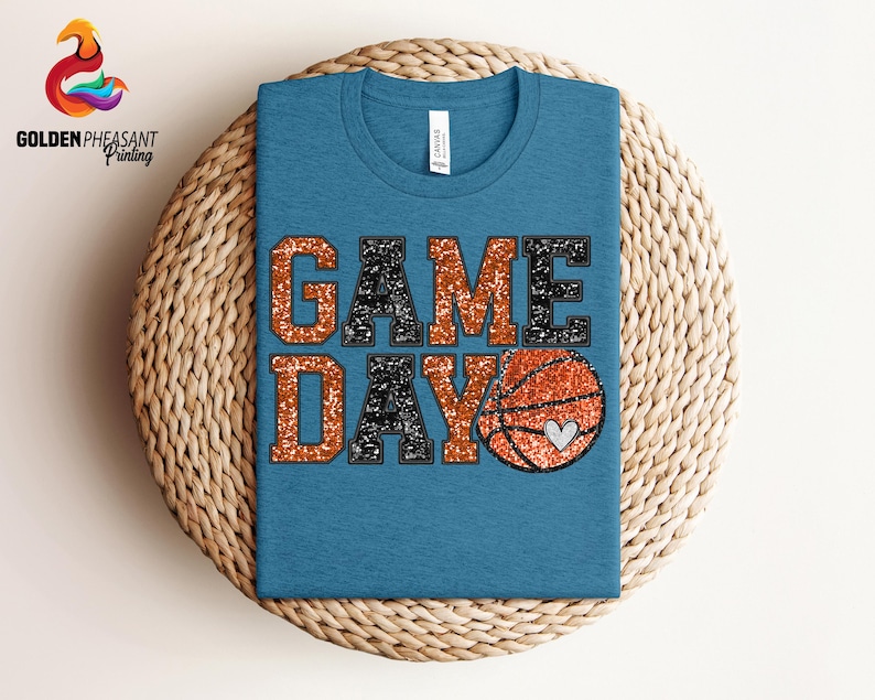 May include: A blue t-shirt with the words "GAME DAY" in black and orange glitter. A basketball with a heart is in the middle of the text.