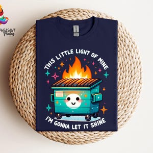 Pu&ograve; includere: Una t-shirt blu navy con un'illustrazione di un cassonetto della spazzatura in fiamme in stile cartone animato. Il testo "This little light of mine" e "I'm gonna let it shine" &egrave; stampato sulla t-shirt.