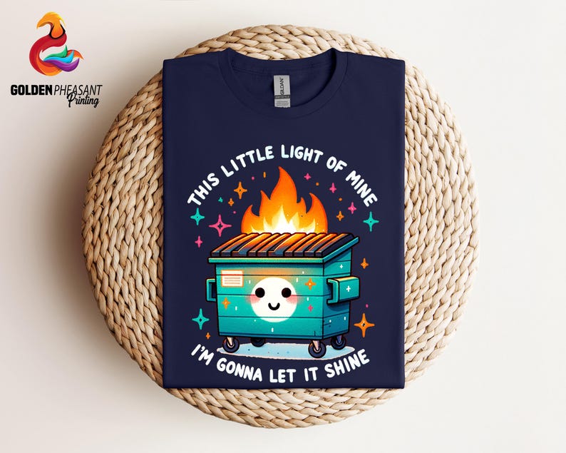 Pu&ograve; includere: Una t-shirt blu navy con un'illustrazione di un cassonetto della spazzatura in fiamme in stile cartone animato. Il testo "This little light of mine" e "I'm gonna let it shine" &egrave; stampato sulla t-shirt.