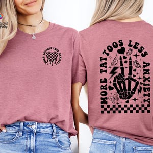 Pu&ograve; includere: Una maglietta rosa con un design grafico nero che dice "Pi&ugrave; tatuaggi, meno ansia" con una mano scheletrica e farfalle.