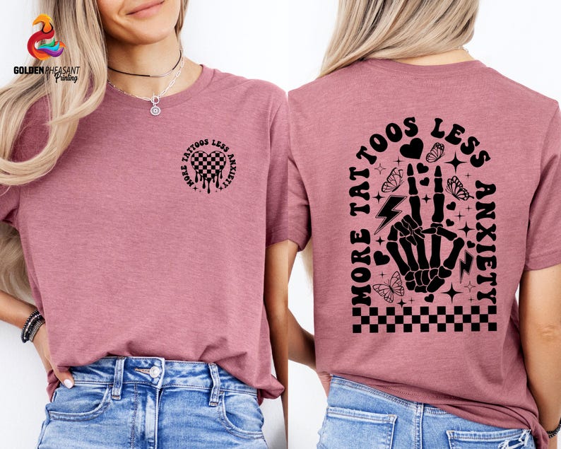 Pu&ograve; includere: Una maglietta rosa con un design grafico nero che dice "Pi&ugrave; tatuaggi, meno ansia" con una mano scheletrica e farfalle.
