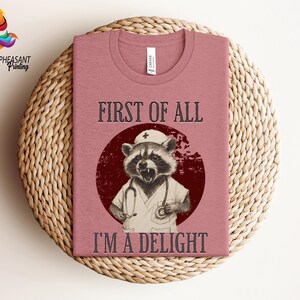 May include: A pink t-shirt with a graphic of a raccoon wearing a nurse's hat and scrubs. The text "First of all, I'm a delight" is printed on the shirt.
