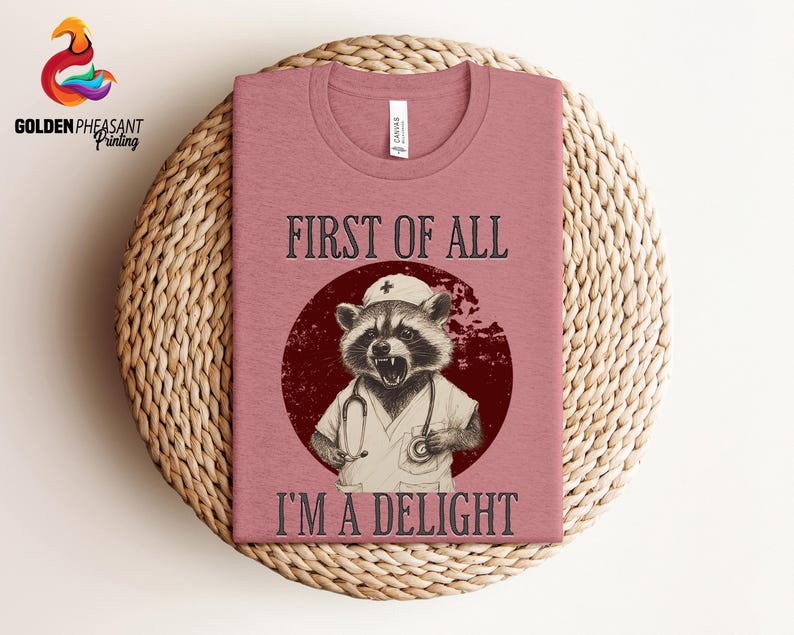May include: A pink t-shirt with a graphic of a raccoon wearing a nurse's hat and scrubs. The text "First of all, I'm a delight" is printed on the shirt.