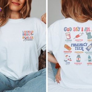 May include: White t-shirt with a colorful graphic design that says "God Say I Am... Pharmacy Tech" with a variety of pills and medical symbols. The text includes bible verses and phrases like "Strong", "Accepted", "Bold", "Redeemed", "Protected", "Courageous", "Kind", "Capable", and "Loyal".