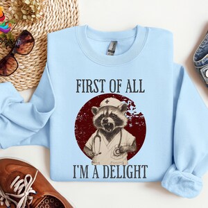 May include: Light blue sweatshirt with a graphic of a raccoon wearing a nurse's hat and holding a stethoscope. The text "First of all I'm a delight" is printed on the sweatshirt.