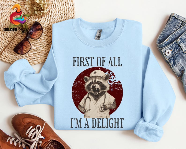 May include: Light blue sweatshirt with a graphic of a raccoon wearing a nurse's hat and holding a stethoscope. The text "First of all I'm a delight" is printed on the sweatshirt.
