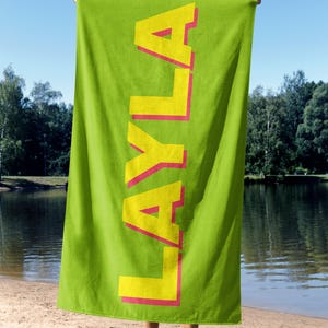 May include: A bright green beach towel with the name "LAYLA" printed vertically in large yellow letters with a pink outline. The towel is held up against a sunny outdoor background, near a lake.