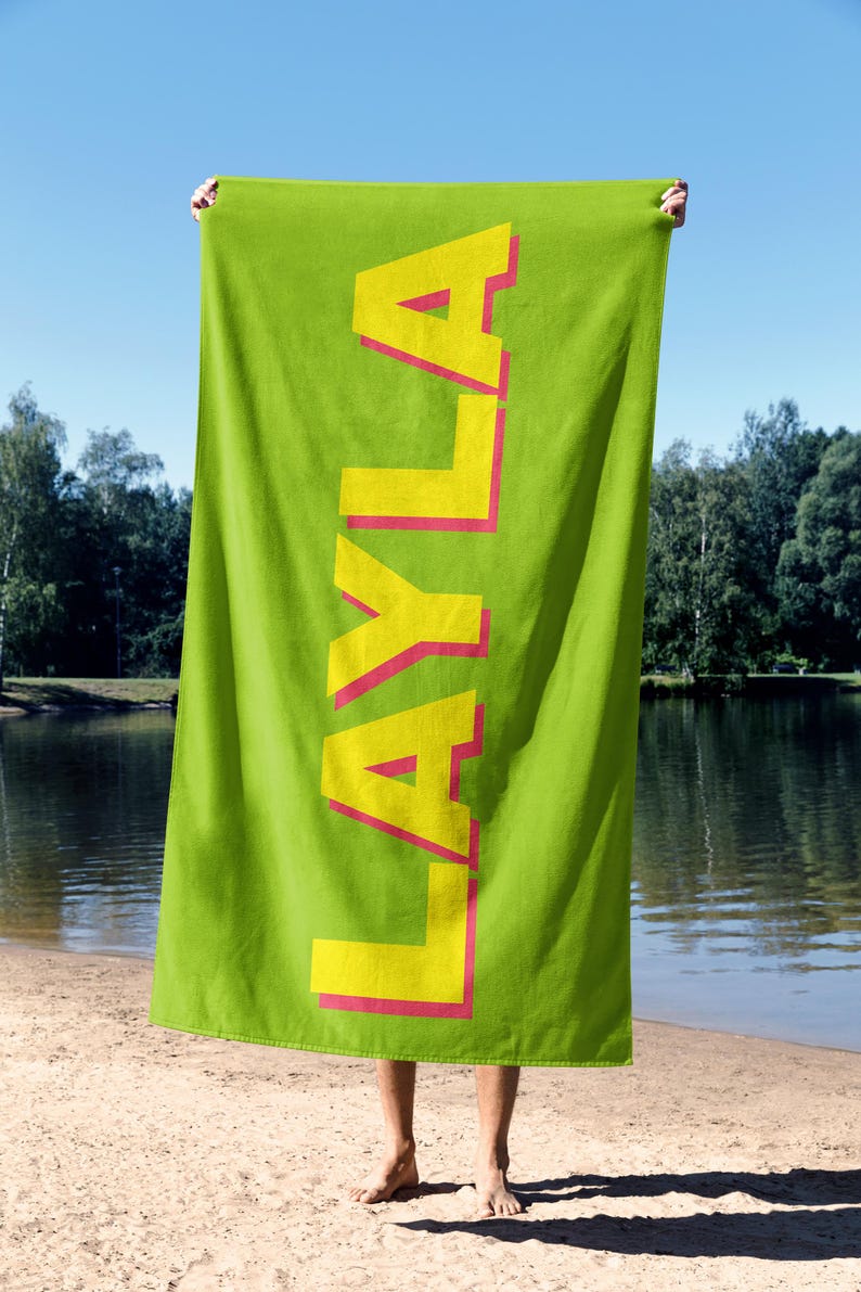 May include: A bright green beach towel with the name "LAYLA" printed vertically in large yellow letters with a pink outline. The towel is held up against a sunny outdoor background, near a lake.