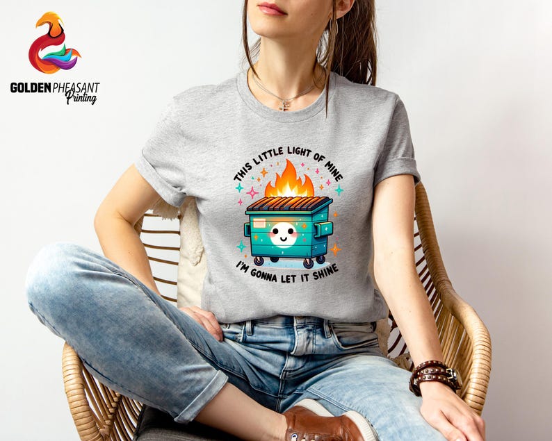 Pu&ograve; includere: Una maglietta grigia con una grafica di un cassonetto verde in fiamme con il testo "This little light of mine, I'm gonna let it shine".