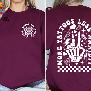 Pu&ograve; includere: Felpa color borgogna con una stampa grafica bianca sul davanti e sul retro. La grafica presenta un cuore con un motivo a scacchi, un design a goccia e il testo "More Tattoos Less Anxiety". La grafica sul retro presenta una mano scheletrica con un fulmine, un motivo a scacchi e il testo "More Tattoos Less Anxiety".