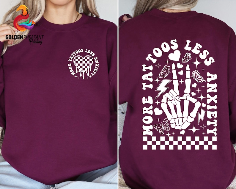 Pu&ograve; includere: Felpa color borgogna con una stampa grafica bianca sul davanti e sul retro. La grafica presenta un cuore con un motivo a scacchi, un design a goccia e il testo "More Tattoos Less Anxiety". La grafica sul retro presenta una mano scheletrica con un fulmine, un motivo a scacchi e il testo "More Tattoos Less Anxiety".