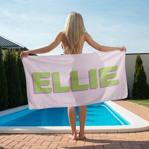 May include: A light pink beach towel with the name "ELLIE" in large, pixelated green letters with a purple outline. The towel is held up in front of a swimming pool on a sunny day.