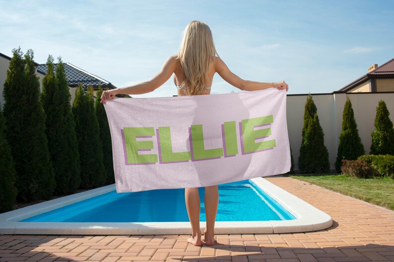 May include: A light pink beach towel with the name "ELLIE" in large, pixelated green letters with a purple outline. The towel is held up in front of a swimming pool on a sunny day.