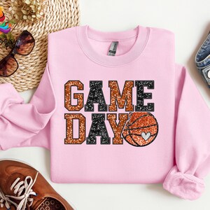 May include: Pink sweatshirt with a glittery orange and black "GAME DAY" graphic and a basketball with a heart on it.