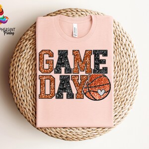 May include: A pink t-shirt with the words "GAME DAY" in black and orange glitter lettering. A basketball with a heart on it is to the right of the text.