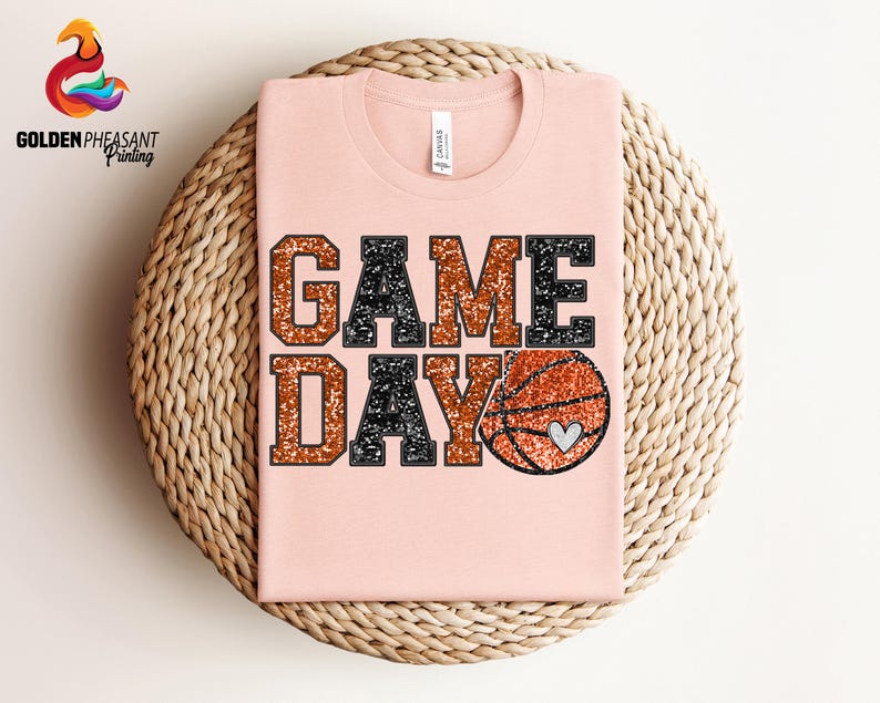 May include: A pink t-shirt with the words "GAME DAY" in black and orange glitter lettering. A basketball with a heart on it is to the right of the text.