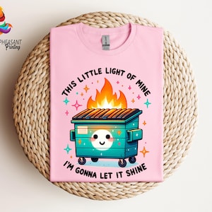 Pu&ograve; includere: Maglietta rosa con una grafica di un cassonetto verde in fiamme con il testo "This little light of mine I'm gonna let it shine".