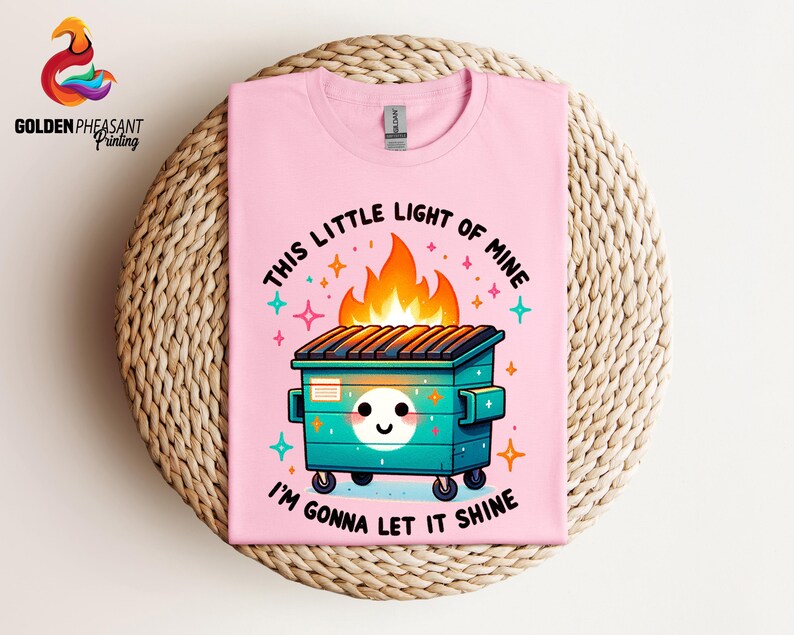 Pu&ograve; includere: Maglietta rosa con una grafica di un cassonetto verde in fiamme con il testo "This little light of mine I'm gonna let it shine".