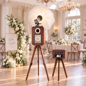 May include: A wooden photo booth station mounted on a tall tripod with a white umbrella light and a ring light. A smaller matching wooden printer stand sits beside it. The screen displays the text Touch here to start in a bright, clear font.