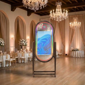 May include: A wooden-framed photo booth with a large, oval-shaped mirror. The mirror has a colorful LED border and displays a blue and white abstract image. The booth is set up in a ballroom with chandeliers and tables.