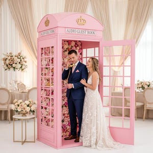 Könnte beinhalten: Eine rosa Telefonzelle mit der Aufschrift "AUDIO GUEST BOOK" oben. Die Zelle ist mit Blumen geschmückt und ein Paar befindet sich darin. Die Frau trägt ein weißes Kleid und der Mann einen blauen Anzug.